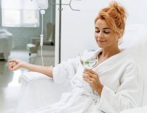 Waist up portrait view of the charming woman in white bathrobe sitting in armchair and receiving IV infusion. She is holding glass of lemon beverage and smiling